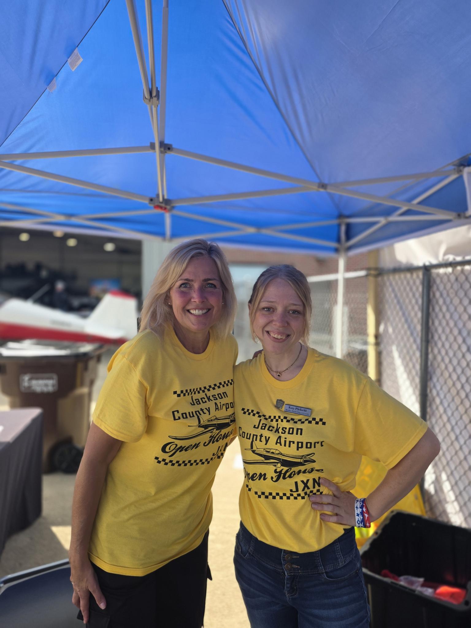Two volunteers in yellow Jackson County Airport Open House shirts smiling together under a blue canopy.