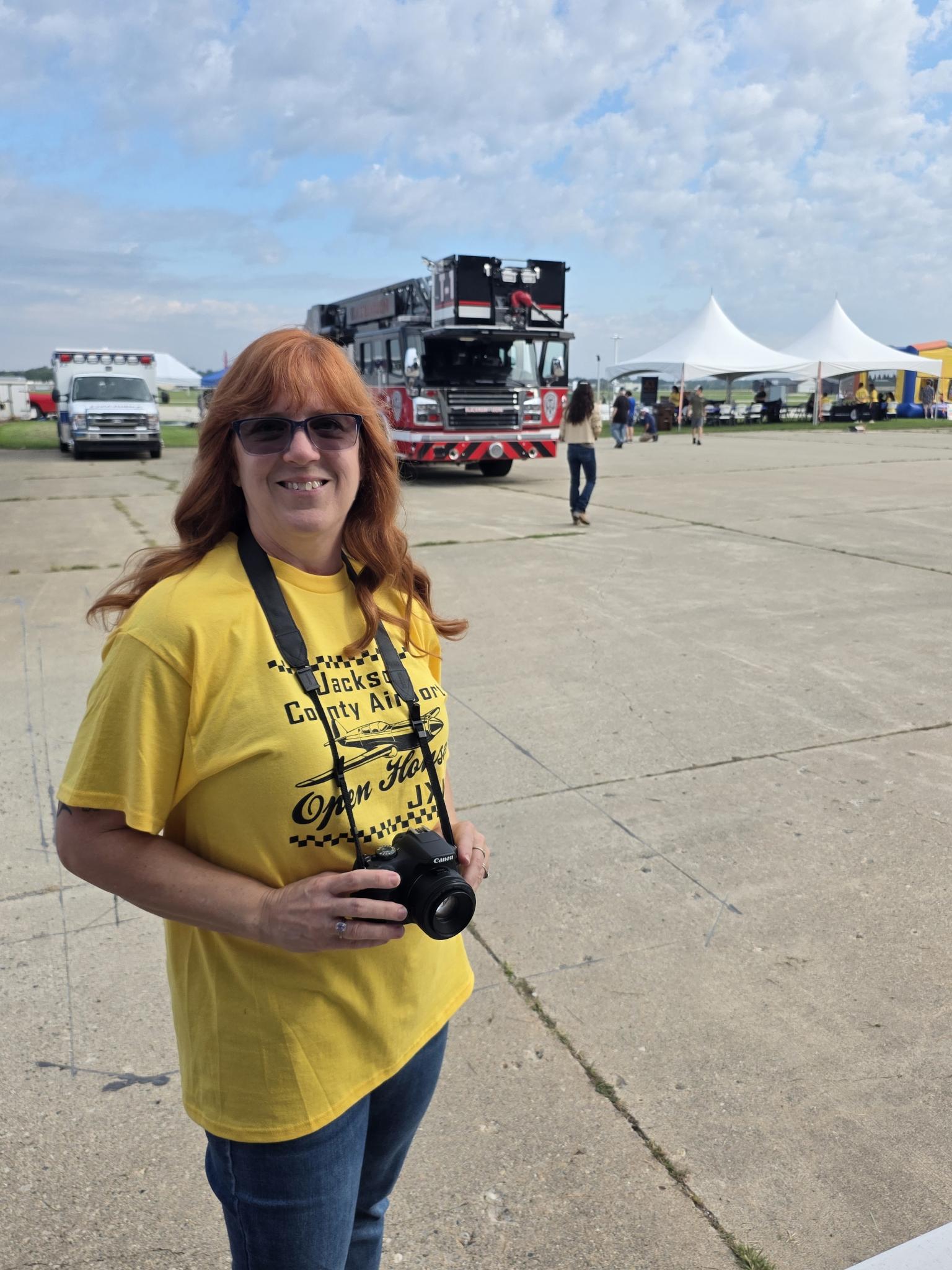 Dawn holding a camera at the Jackson County Airport Open House, wearing a yellow volunteer shirt.