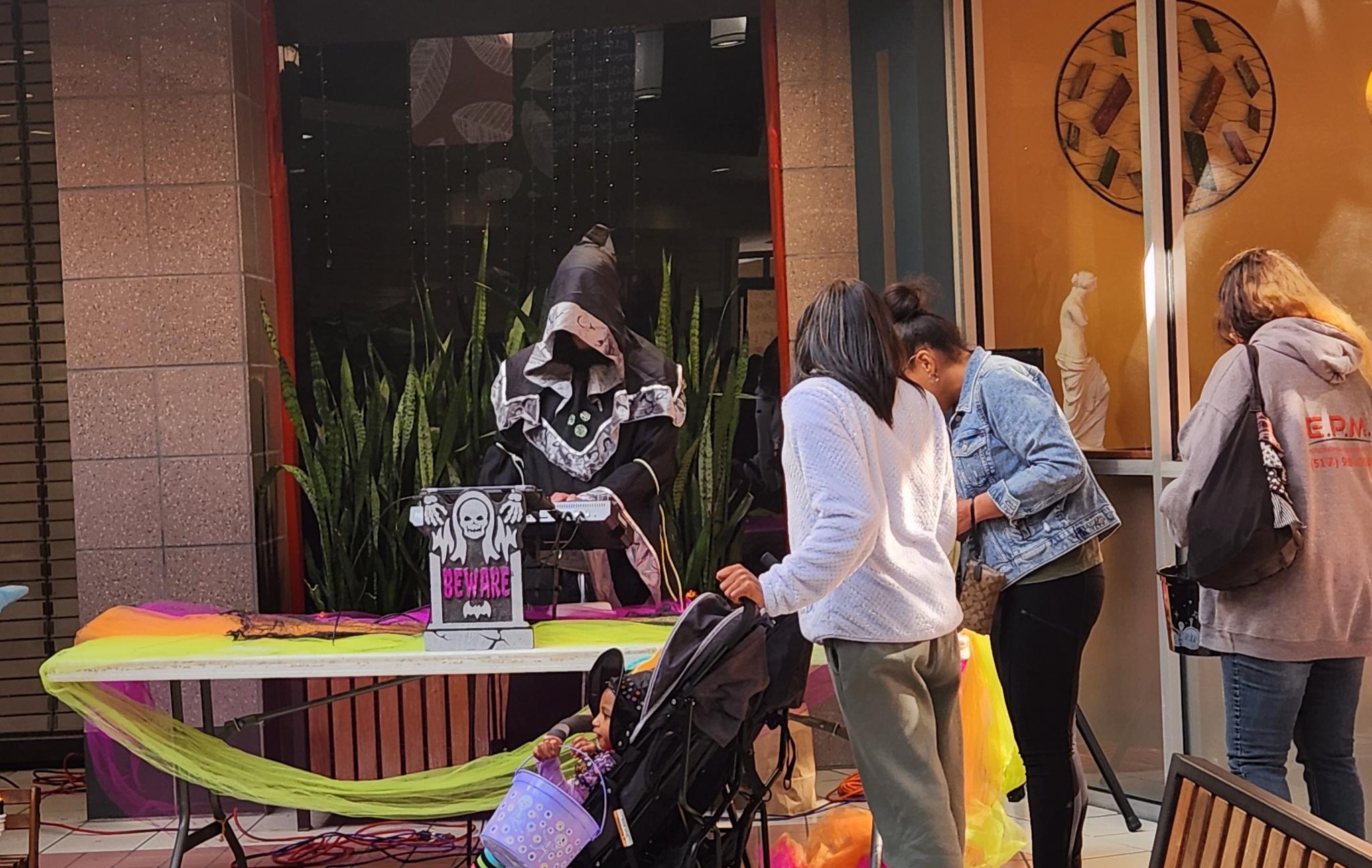 Visitors gathering around a costumed DJ at a Halloween-decorated booth during Malloween.