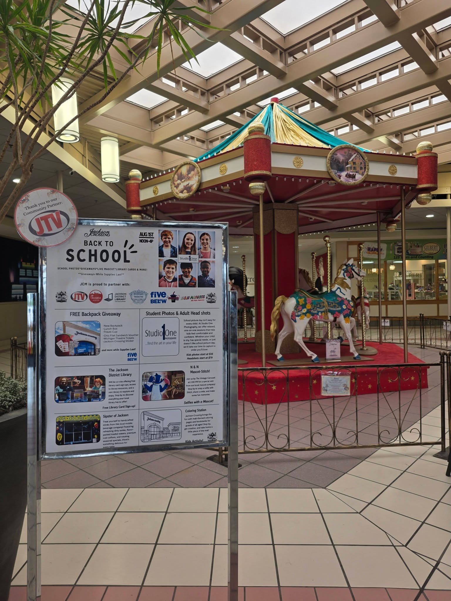 Back to School Bash information board beside the Jackson Crossing carousel, listing backpacks, school photos, library sign-ups, and activities.